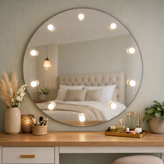 Apvalus veidrodis su integruotomis LED lemputėmis virš kosmetinio staliuko jaukiame miegamojo interjere su dekoratyvinėmis gėlėmis ir aksesuarais.